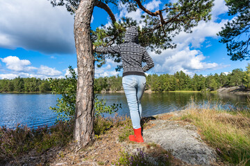 Alone beautiful girl on the forest coast of Scandinavia on fall sunny day. Young slim woman female traveler dressed in hoodie and blue jeans relaxing watching panorama lake water in autumn.