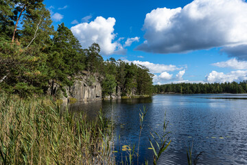 Fototapeta na wymiar Beautiful lake high rocky stony shore on sunny autumn day. Water lily leaves cover the surface of water. Fall season landscape. Blue sky white clouds. Colorful trees on the shore. Scandinavia views.