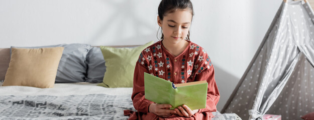 preteen girl reading book while sitting on bed near pillows and wigwam, banner.