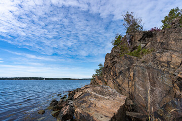 Young slender woman climbs up the rocks. Sports girl dressed in a hoodie and leggings climbs without insurance on high rock on the seashore. Panoramic view of skyline horizon and white clouds over.