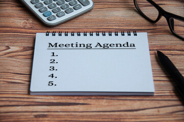 Meeting agenda text on notepad with calculator, pen, glasses and wooden table background.