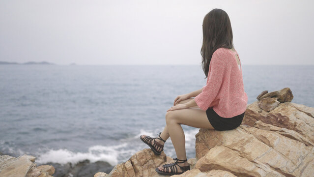 A beautiful woman in pink sweater sits on a rock above the sea, looking at the ocean. Girl traveler, backdrop of the sea and sky. thinking, freedom, solitude, relaxed, Enjoying concept. copy space.