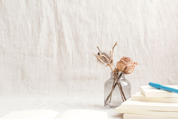 ピンクのバラのドライフラワーが入った花瓶とペンとノートとテクスチャのあるグレーの布の空間	
