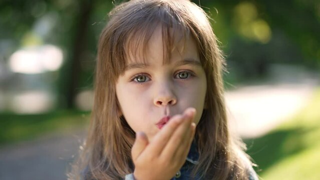 Close-up pretty little girl gesturing air kiss in slow motion looking at camera. Front view headshot of cute Caucasian kid posing in summer spring park enjoying weekend leisure. Lifestyle concept