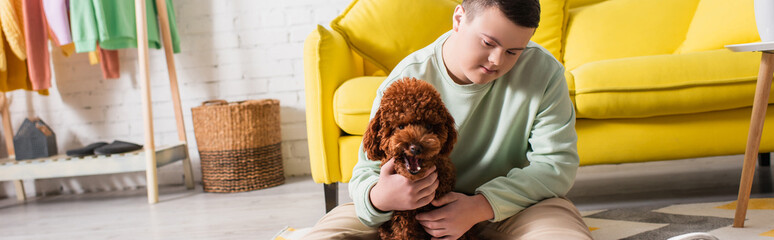 Teenage boy with down syndrome petting brown poodle on floor at home, banner.