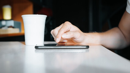 Close-up of male caucasian hand using smartphone in morning indoors. Side view of hand touching screen of mobile phone and mug of coffee standing on table. Selective focus on phone