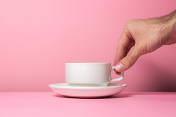 A man's hand holds a white cup on a pink background. Lift the cup from the saucer