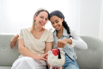 Obraz na płótnie Canvas Adorable black teen daughter with mother sit on sofa at home with piggy bank