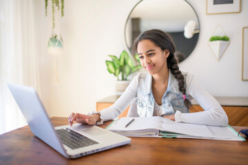 African American teen girl learning school tasks at home, writing notes and using laptop on the...