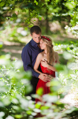 Young beautiful lovers guy and girl in a red dress are hugging in the summer in a green forest