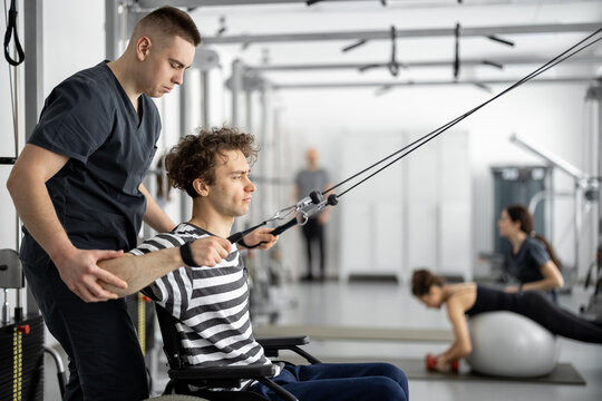 Rehabilitation specialist helps a guy in a wheelchair to do exercise on decompression simulator for recovery from injury. Concept of physical therapy for people with disabilities