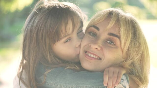 Close-up portrait of smiling Caucasian woman looking at camera as girl kissing cheek in slow motion. Happy young beautiful mother posing in sunshine with cute little daughter. Family leisure
