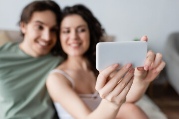 blurred curly woman and happy man taking selfie in bedroom.