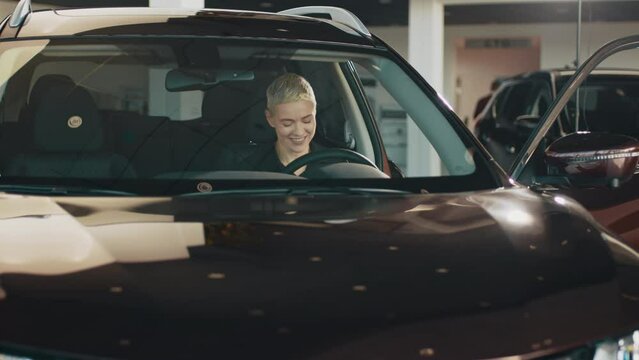 Woman driving a new car. Woman Driver Portrait at Car. Woman smiling in her new car in a showroom. Woman driving a new luxury electric car in a dealership. The girl shows emotions