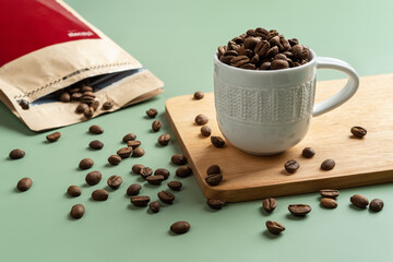 Roasted coffee beans in a small white cup on a pastel green background. Whole coffee beans spilled...