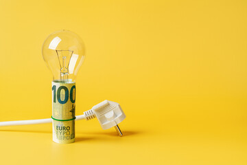 Light bulb on a rolled 100 euro banknotes and white power plug over yellow background. Copy space....