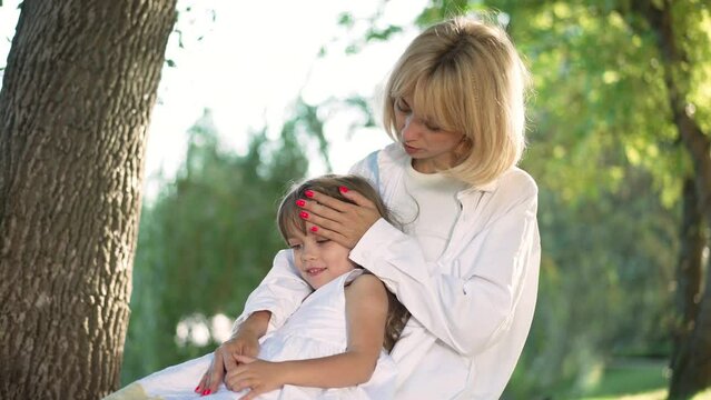 Happy young mother hugging daughter stroking hair in slow motion talking. Portrait of smiling confident Caucasian woman enjoying leisure in summer spring park with pretty charming girl