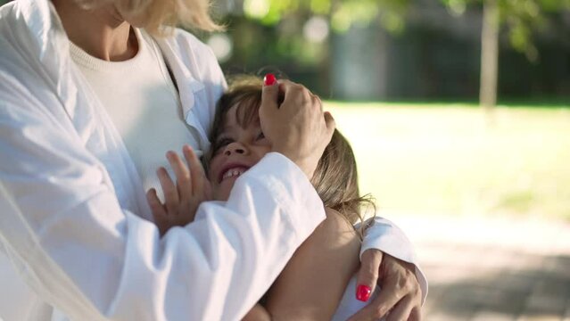 Beautiful little girl admiring unrecognizable woman sitting in sunny park outdoors. Portrait of happy relaxed Caucasian daughter hugging mother enjoying leisure with parent outdoors. Slow motion