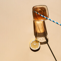 Glass of beverage with lemon slice and straw on hot summer’s day. Simple square flat lay composition with long shadows and light reflections.