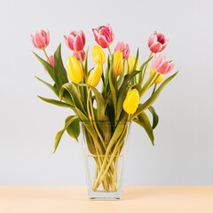 Bouquet of yellow and pink tulip flowers in transparent glass vase on sand coloured table. Simple composition on light pastel blue background.