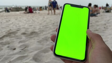 Using phone with green screen vertical chroma key close up man hand swiping, scrolling, tapping, pinching on screen. Blurred crowd tourist walking, relaxing, take selfie on beach in background - Powered by Adobe