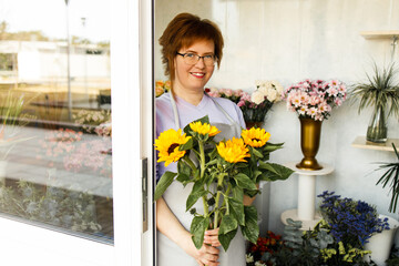 Woman florist standing and holding bouquet of sunflowers in flower shop. Female in apron holds bouquet of flowers for client and making a bouquet of fresh flowers