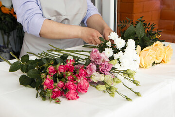 Fototapeta na wymiar Startup, small business, flower shop. Female in apron holds bouquet of flowers for client and making a bouquet at front door of plants studio. Close up of beautiful flowers lying on table