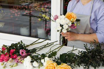 Startup, small business, flower shop. Female in apron holds bouquet of flowers for client and making a bouquet at front door of plants studio. Close up of beautiful flowers lying on table