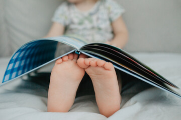 little girl holding bare feet close up to camera and reading book . Blurred face on background 