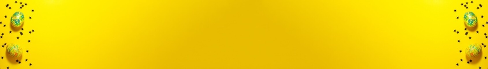 Banner of two green and yellow easter eggs between dark stars on the yellow background on the left and right. Flat lay with copy space. Holidays concept