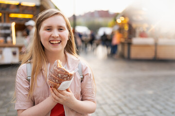 Portrait of happy modern girl at fair in city eating trdelnik