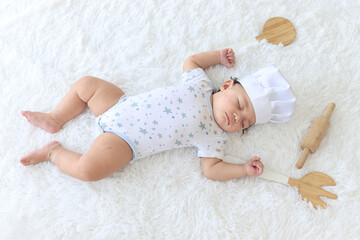 Six months crawling baby kid sleeping next to kitchenware and wooden rolling pin on fluffy white rug, wearing chef hat, costumes like a chef, childhood and dreaming career concept.