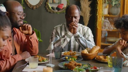 African American family praying with eyes closed before meal while sitting together at dinner table at home