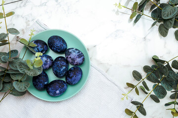 Cosmic-colored eggs lie in a green plate, next to a white waffle towel on a marble table with green leaves. Easter