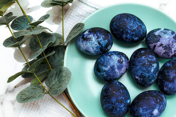 Cosmic-colored eggs lie in a green plate, next to a white waffle towel on a marble table with green leaves. Easter