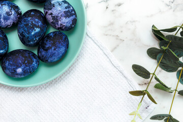 Cosmic-colored eggs lie in a green plate, next to a white waffle towel on a marble table with green leaves. Easter