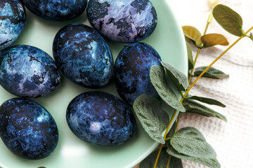 Cosmic-colored eggs lie in a green plate, next to a white waffle towel on a marble table with green leaves. Easter
