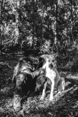 Happy, young, caucasian and handsome guy with jacket and leather shoes petting a beautiful white and brown dog in the ground of the forest, Valdivia, Chile (in black and white)