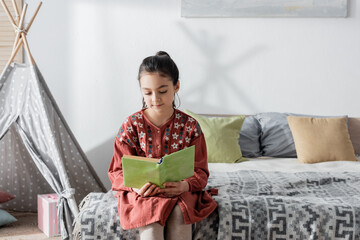 preteen girl reading book while sitting on bed near pillows and wigwam on background. © LIGHTFIELD STUDIOS