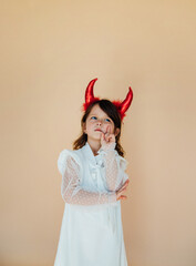 Portrait of a little girl in a white dress with devil horns. Halloween costume. Angelic facial...