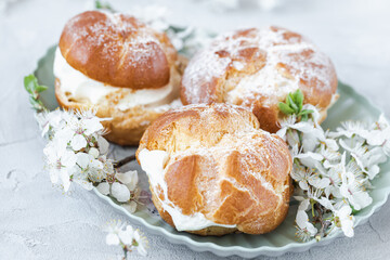 Choux Buns with whipped cream and sugar powder on top on plate. Gray background with spring white cherry blossom flowers. Delicate choux pastry dessert. French cream puffs