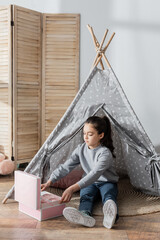 full length view of girl sitting in wigwam near box with toy tea set. © LIGHTFIELD STUDIOS