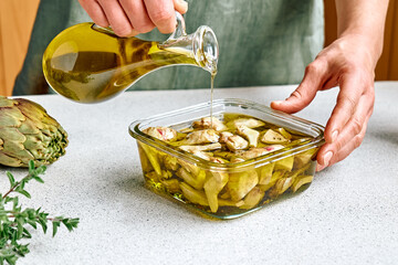 Artichoke hearts marinated with olive oil and herbs. Woman pouring oil in glass jar with pickled...