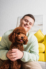 Teen boy with down syndrome hugging poodle and looking at camera in living room.