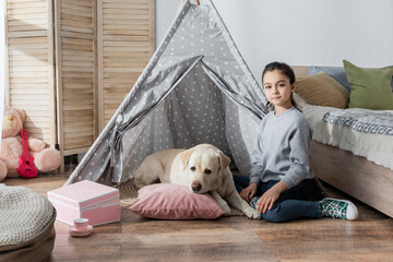 preteen girl looking at camera near labrador lying on pillow in wigwam.