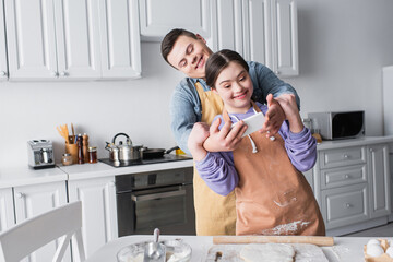 Cheerful couple with down syndrome taking selfie on smartphone near dough in kitchen.