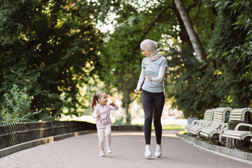 Fototapeta premium Family sports and outdoor activity concept. Cheerful young Muslim Arabian woman enjoying running together in park with her cute active 4-aged daughter. Mom and child jogging in the park