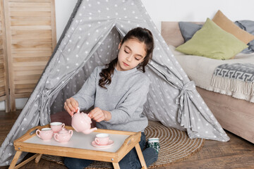 preteen girl sitting in wigwam and pouring tea from toy teapot. © LIGHTFIELD STUDIOS