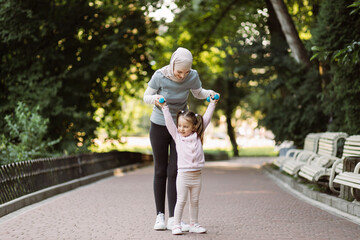 Beautiful young muslim arab woman and her charming little daughter are smiling while working out with dumbbells outdoors in the park. Mom showing her child how to raise dumbbells
