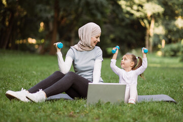 Full length of Muslim mother and little daughter sitting on yoga mats outdoors in the park. Smiling mom in hijab showing her kid how to exercise with dumbbells, watching training video on laptop
